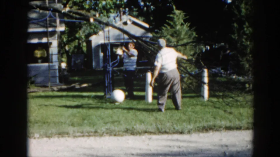 Man viewed from behind in a suburban Illinois yard, c. 1962, with fallen tree branch arching overhead and single-story house behind; 8mm home movie footage.