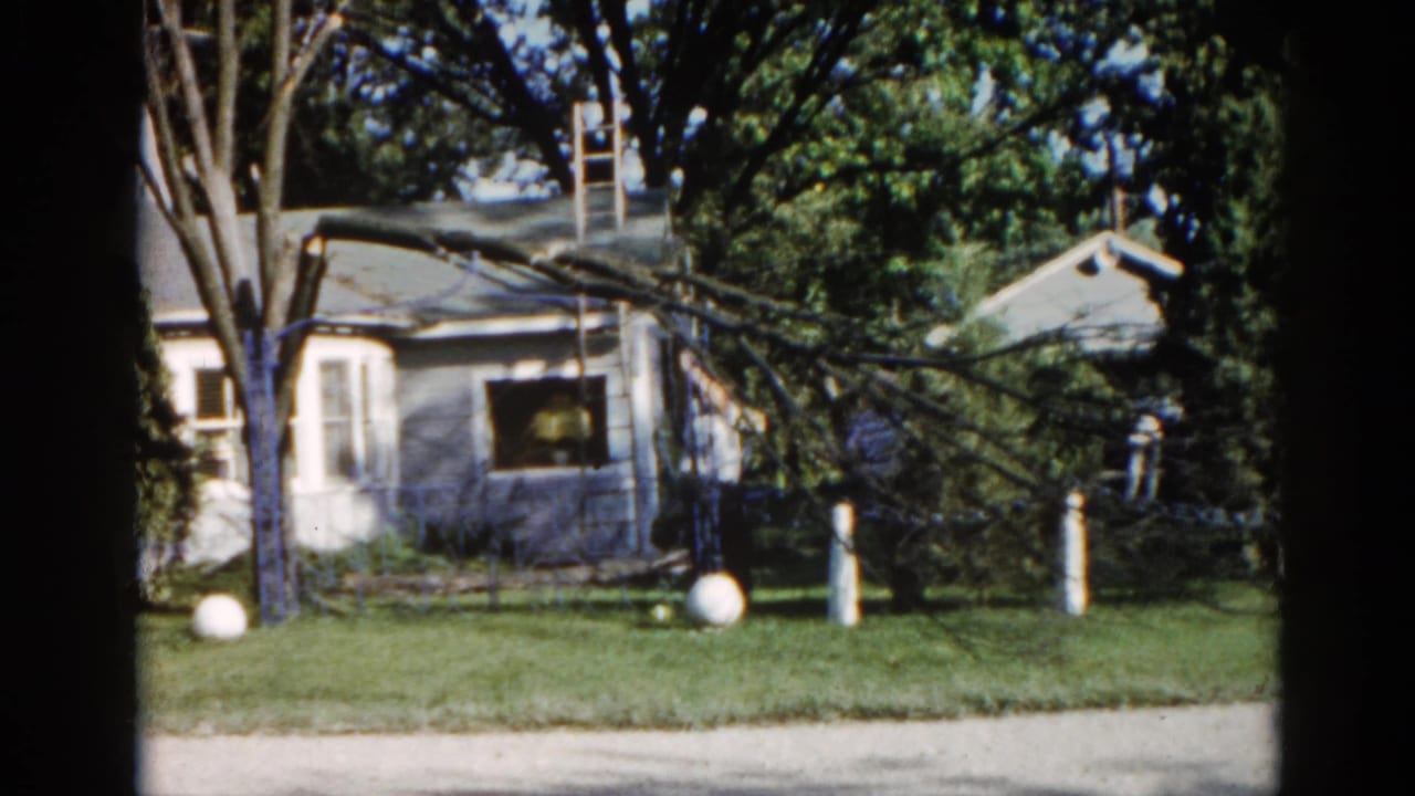 8mm home movie frame from 1962 Illinois showing a ranch-style suburban house, fallen tree branch, ladder against the roofline, and two white spherical lawn ornaments.