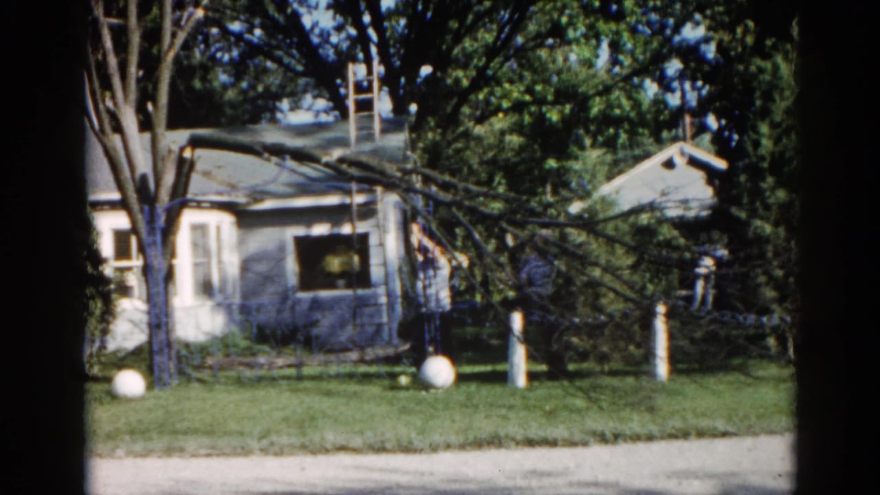 Second frame from a 1962 Illinois 8mm home movie showing the same ranch house, fallen branch, ladder, and a distant figure near the eaves.