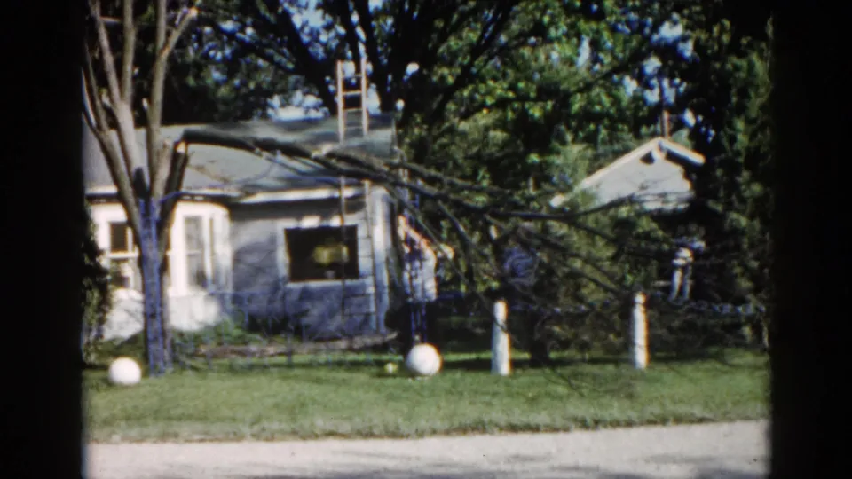 Second frame from a 1962 Illinois 8mm home movie showing the same ranch house, fallen branch, ladder, and a distant figure near the eaves.