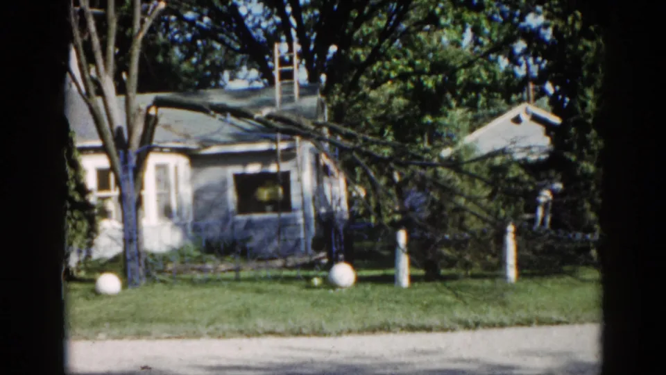 Mid-clip frame from 1962 Illinois 8mm footage showing the ranch house with fallen storm branch, ladder against roofline, and white spherical lawn ornaments on the grass.