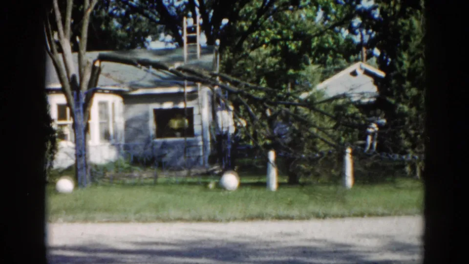 Closing frame from a 1962 Illinois 8mm home movie showing a ranch house with a fallen tree branch, a ladder propped against the roof, and white spherical ornaments on the lawn.