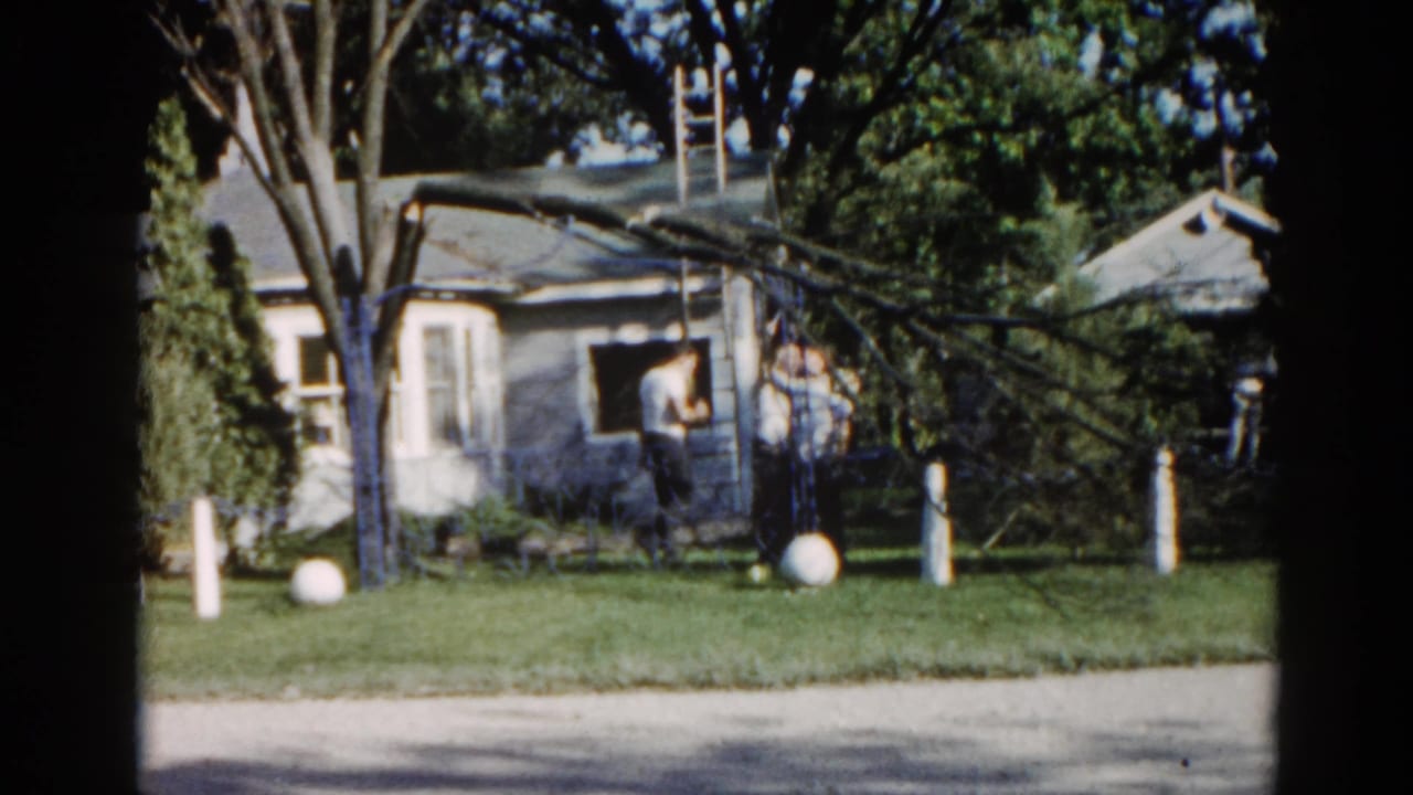 1962 Illinois: men with a ladder working to remove a storm-damaged tree limb in a suburban yard beside a mid-century ranch house.