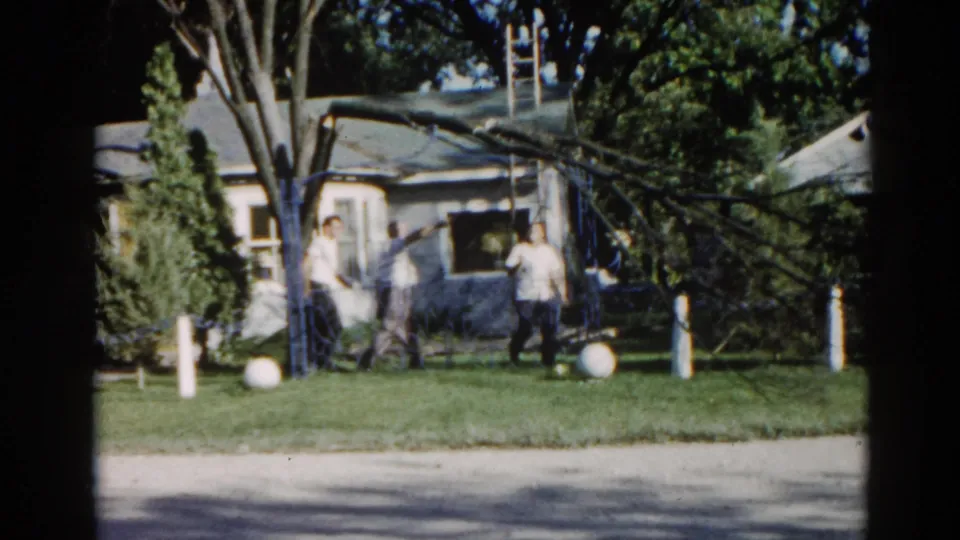 8mm home movie, c.1962, Illinois: a man in a light shirt surveys a suburban yard with a large fallen tree resting against the grey ranch-style house behind him.