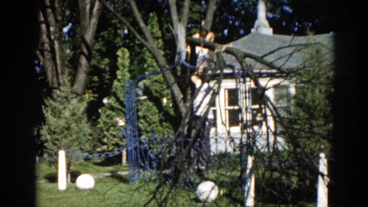 8mm home movie frame from c. 1962 Illinois showing a white clapboard house, ornamental blue fence, and white globe lawn ornaments in a suburban backyard.