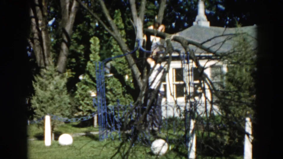8mm home movie frame from c. 1962 Illinois showing a white clapboard house, ornamental blue fence, and white globe lawn ornaments in a suburban backyard.