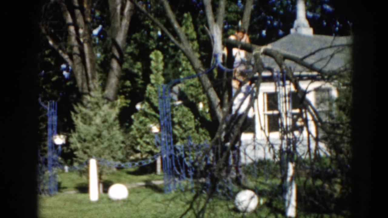 Near-identical view of white house with ornamental top, blue decorative fencing, and white spherical lawn ornaments amid leafy trees.