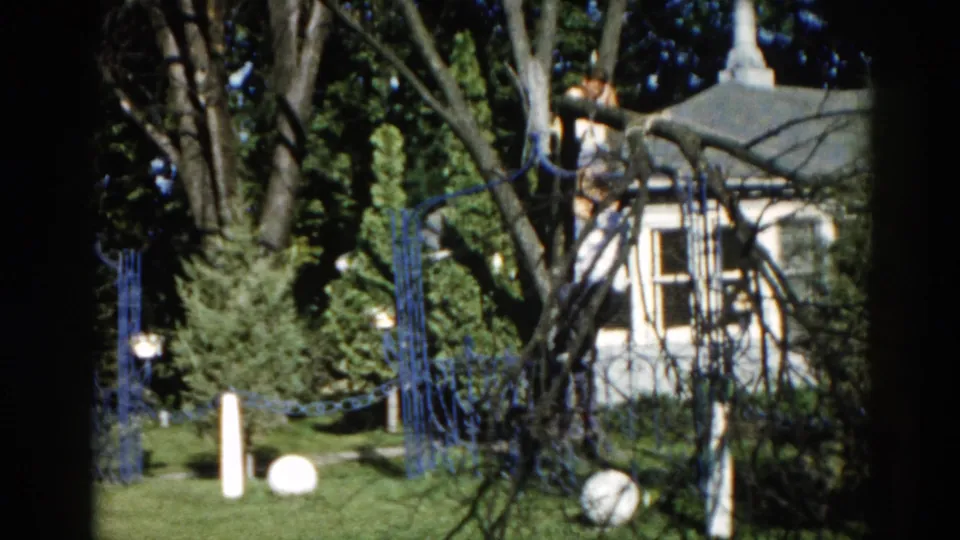 Continuation 8mm frame from c. 1962 Illinois suburban backyard showing the white house, ornamental blue fence, and globe lawn decorations with slight camera movement.