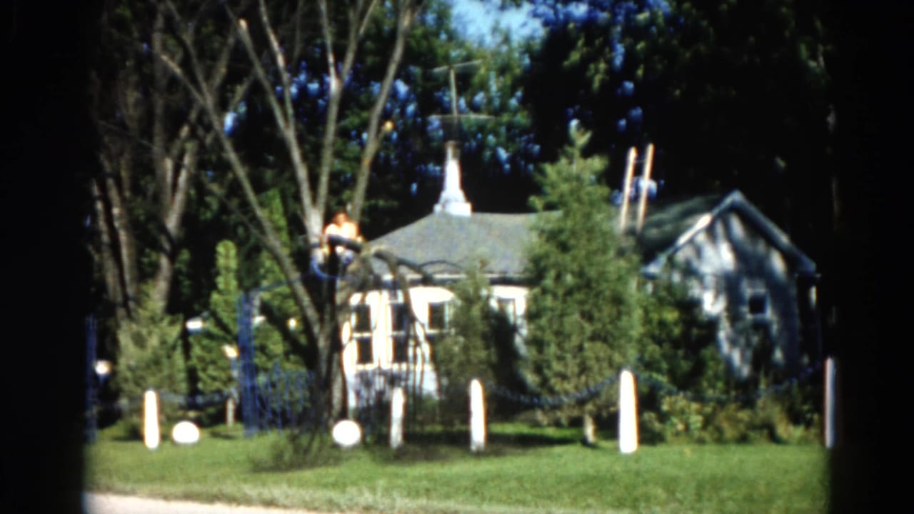 Wide 8mm shot of a suburban yard with a man high in a tree, a peaked-roof house, white fence posts, and blue fence rails in natural daylight.