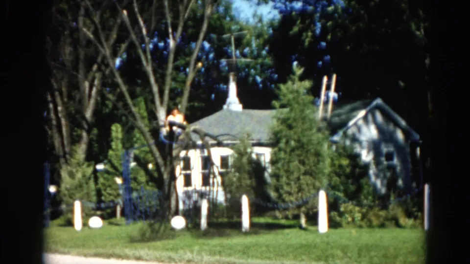 8mm home movie frame from 1962 Illinois showing a man working in a tall tree above a suburban house with a distinctive cupola-style peaked roof.