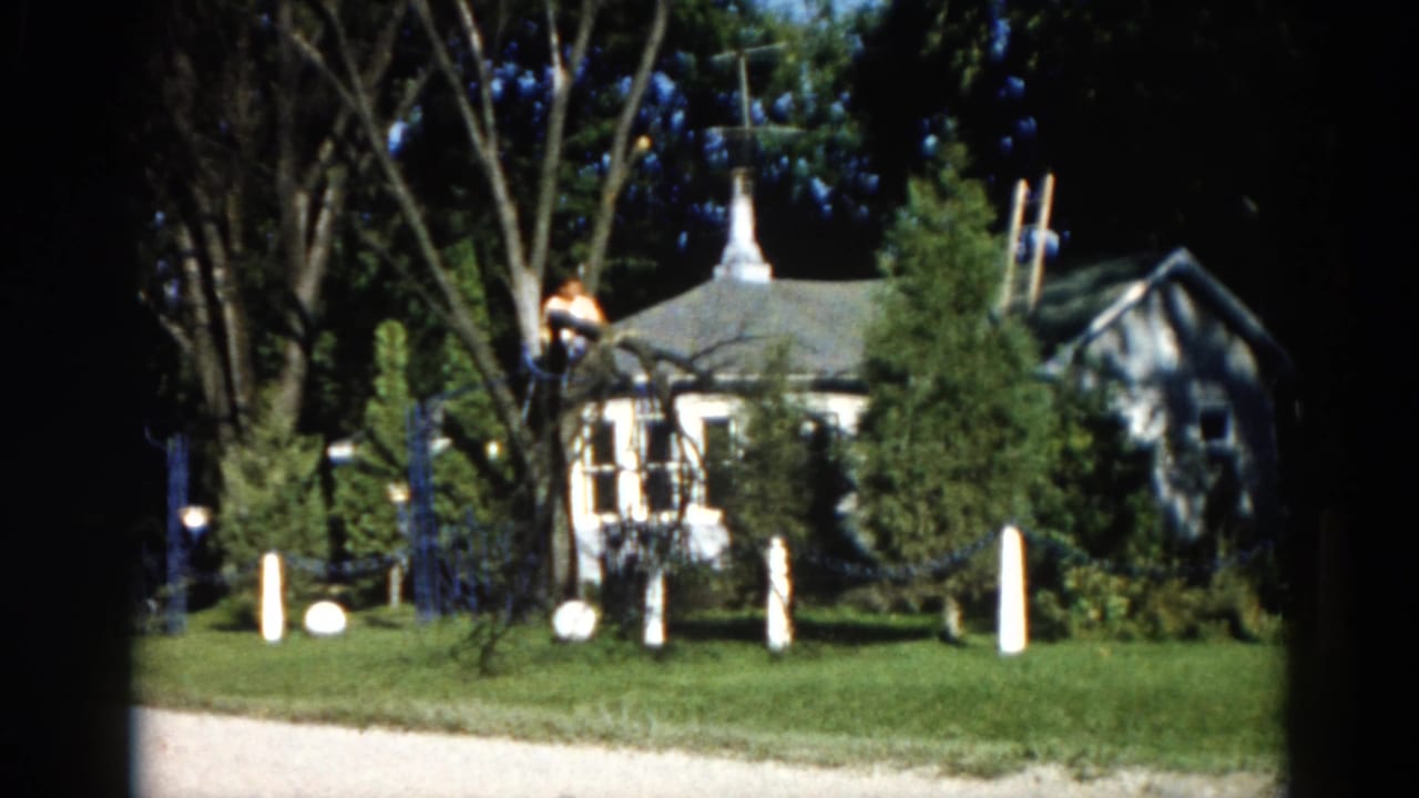 1962 Illinois 8mm frame showing a man in a tall tree with a suburban house featuring a steeple-like peaked roof visible behind leafy canopy.