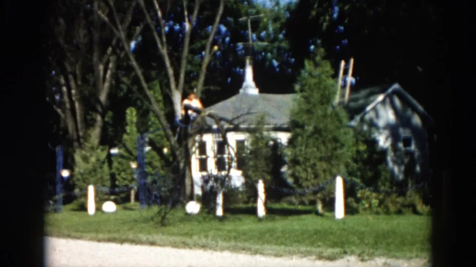 1962 Illinois 8mm frame showing a man in a tall tree with a suburban house featuring a steeple-like peaked roof visible behind leafy canopy.