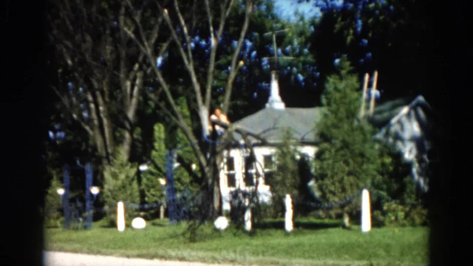 Mid-action 8mm frame from 1962 Illinois; man visible in upper tree branches above a suburban house with a distinctive steeple-like roofline.