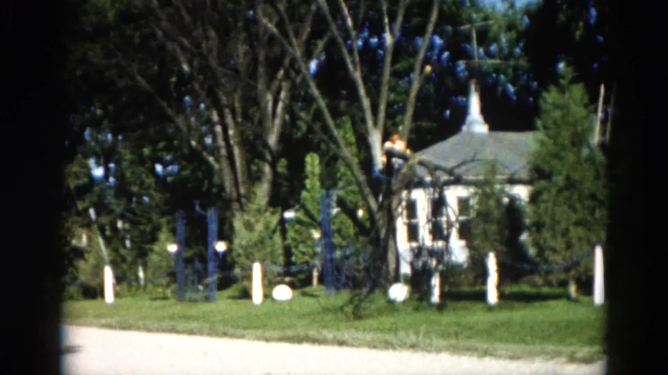 Final 1962 Illinois 8mm frame showing man in tree above suburban house; a light-coloured driveway and decorative fence posts appear in the foreground.
