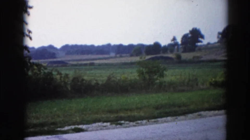 8mm home movie frame, 1962 rural Illinois — open farmland with a low rounded mound in the middle distance, tree-lined horizon, and pale overcast sky.