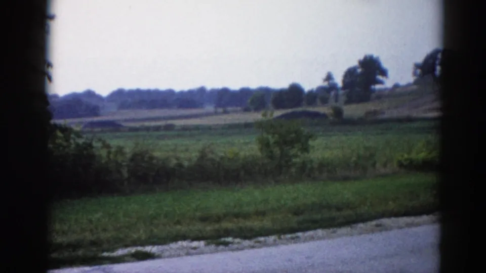 Closing frame of a 1962 Illinois home movie drive — countryside slides past a car window, green fields and a tree-lined horizon receding under a pale grey sky.