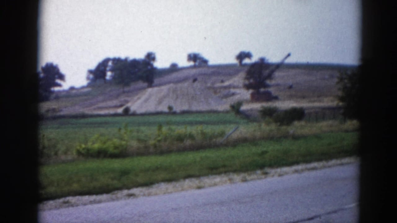 8mm home movie from 1962 Illinois — green fields, a large earthwork on a hill, and a crane arm silhouette, framed through a car window.