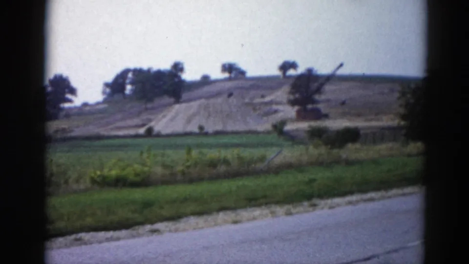 8mm home movie from 1962 Illinois — green fields, a large earthwork on a hill, and a crane arm silhouette, framed through a car window.