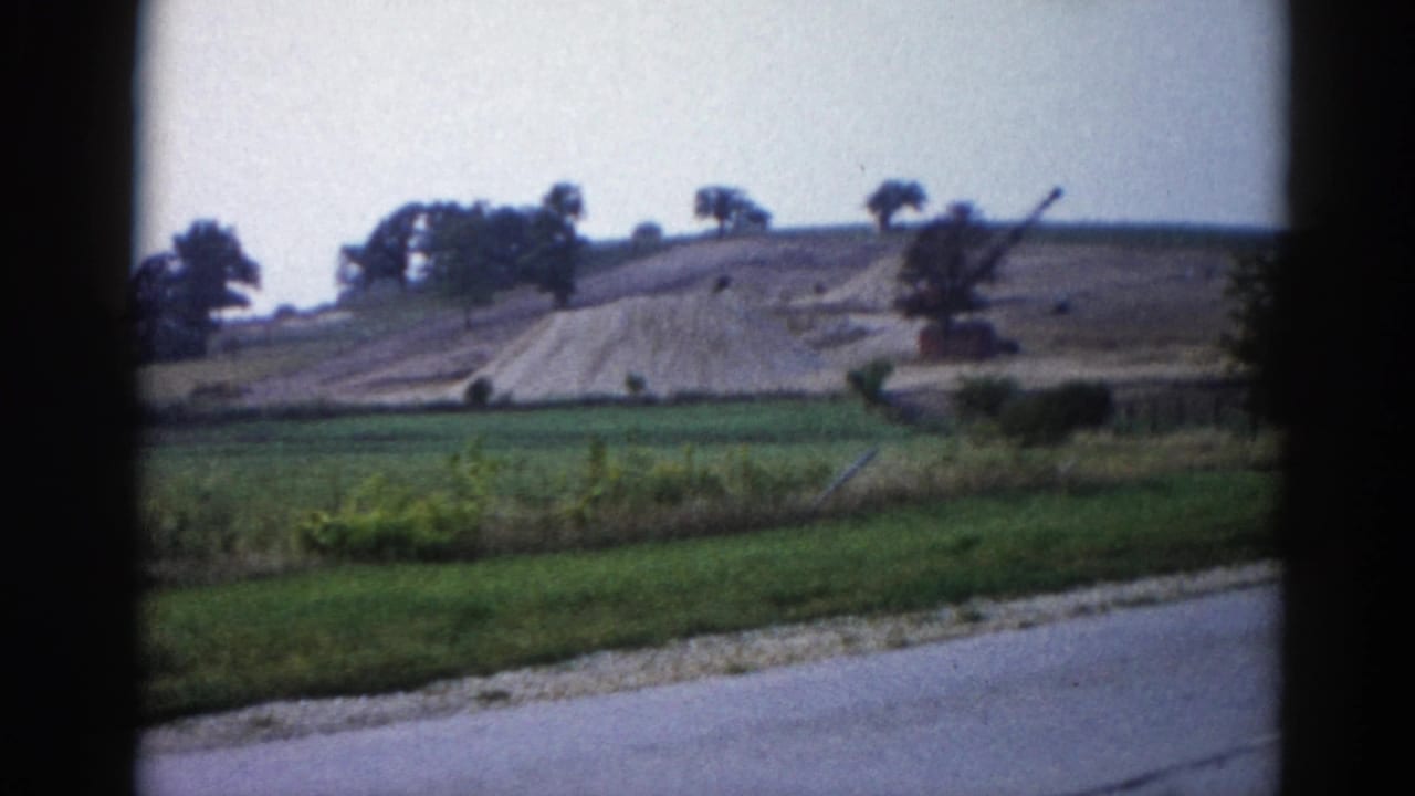 8mm frame from 1962 Illinois road trip — vehicle still passing the earthwork; graded hill and equipment boom visible above green pasture.
