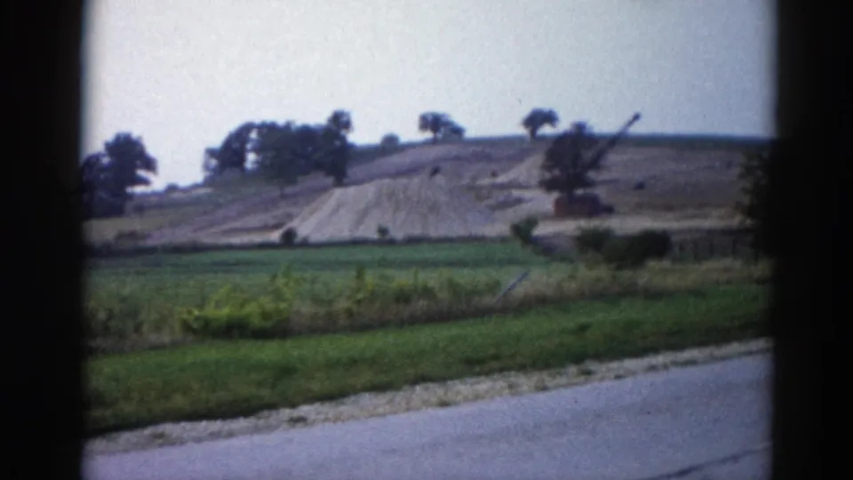 8mm frame from 1962 Illinois road trip — vehicle still passing the earthwork; graded hill and equipment boom visible above green pasture.