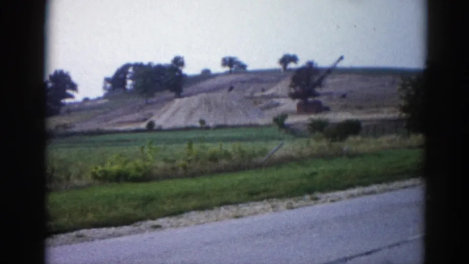 Mid-sequence 8mm frame from 1962 Illinois — roadside shrubs visible at right, earthwork and crane arm still on distant hill above green fields.