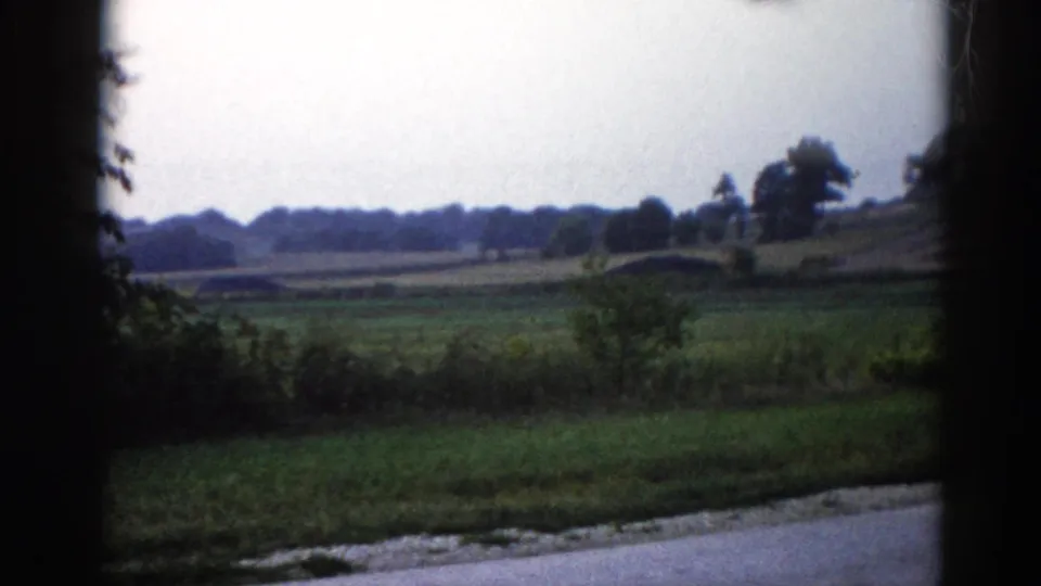 Final 8mm frame from 1962 Illinois — construction site now out of view; open rural landscape, low treeline, roadside shrubs, and pale flattened sky.