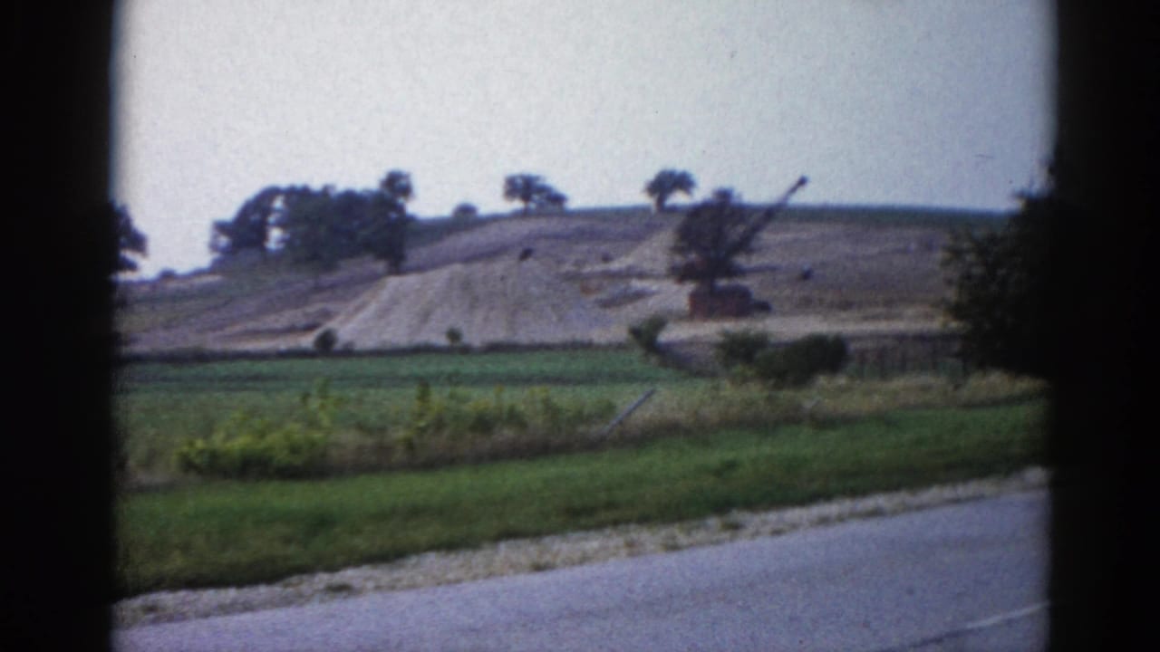 Earthen hillside under construction with a crane visible at right, green fields and a paved road in foreground.