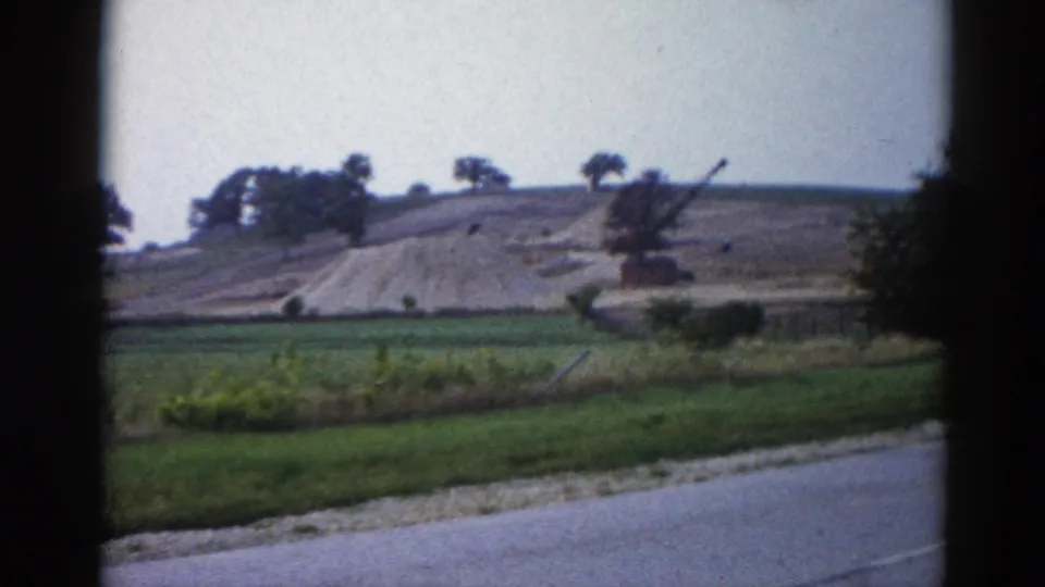 Graded earthen embankment with construction crane at right, scattered trees, green grass and road edge visible, shot on 8mm film in Illinois circa 1962.