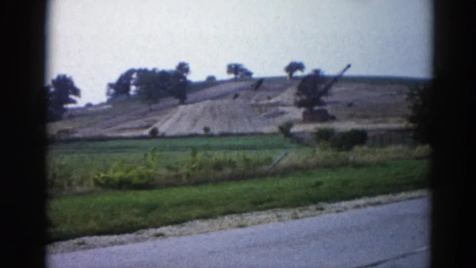 Continuing shot of the graded hillside and crane from a vehicle, green roadside fields and paved road in foreground, 8mm footage from rural Illinois circa 1962.