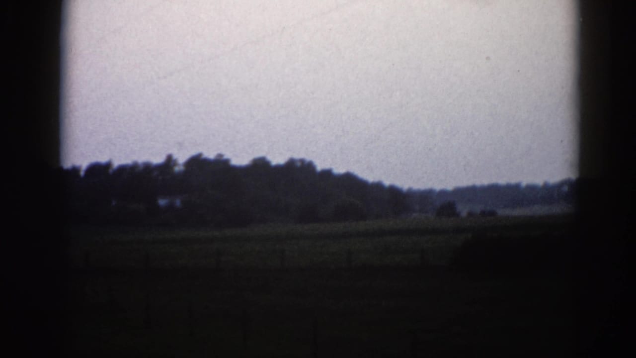 8mm home movie frame from Illinois, c. 1962 — flat field, low tree-line horizon, and overcast sky with heavy dark film-edge vignetting.