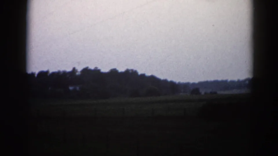 8mm home movie frame from Illinois, c. 1962 — flat field, low tree-line horizon, and overcast sky with heavy dark film-edge vignetting.