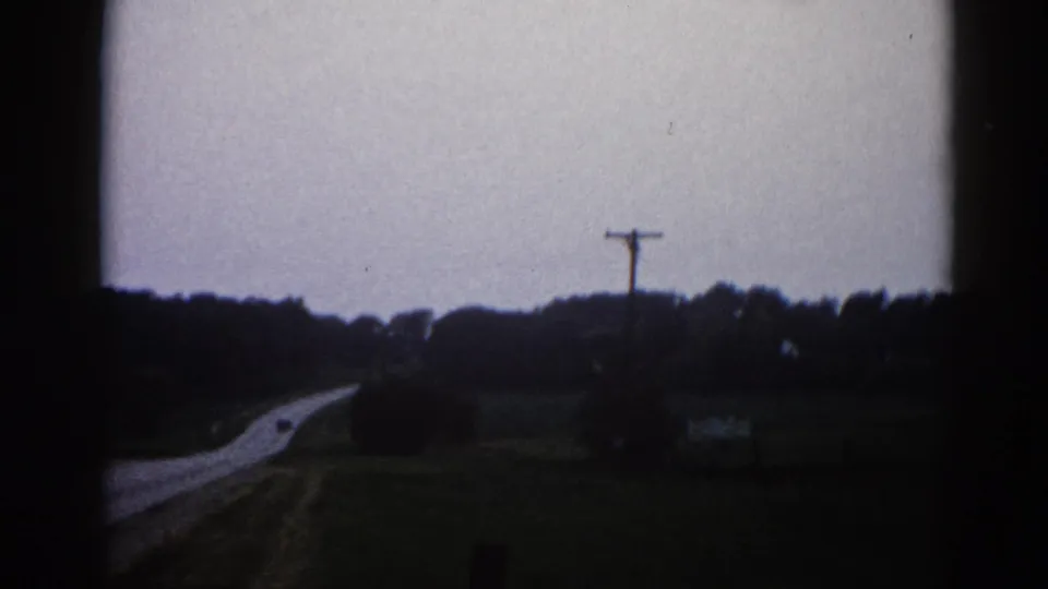 Roadside panoramic view from a moving vehicle in rural Illinois, 1962 — two-lane road, utility pole, flat fields, and overcast sky on 8mm film.