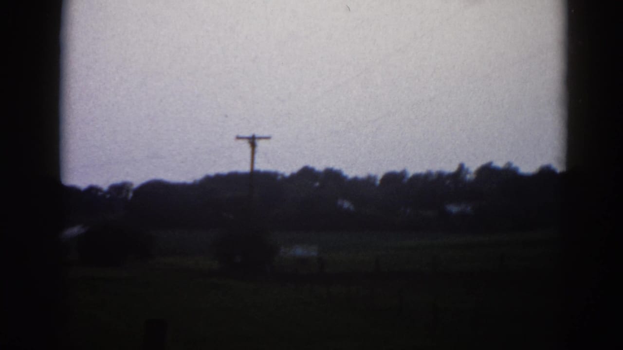 Continuing roadside pan in rural Illinois, 1962 — utility pole centered in frame, treeline, low farm buildings, and overcast sky on 8mm film.