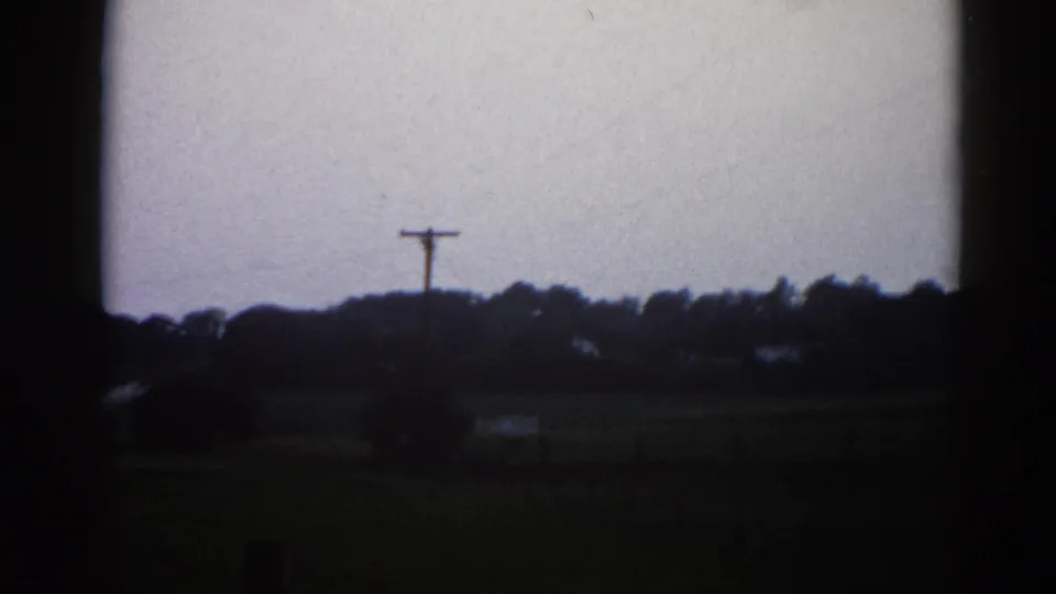 Continuing roadside pan in rural Illinois, 1962 — utility pole centered in frame, treeline, low farm buildings, and overcast sky on 8mm film.
