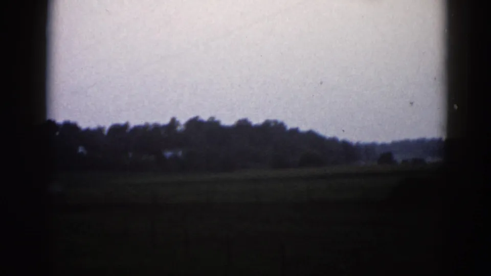 Final frame of the roadside pan in rural Illinois, 1962 — open fields, distant treeline, low farm buildings, and overcast gray sky on 8mm film.