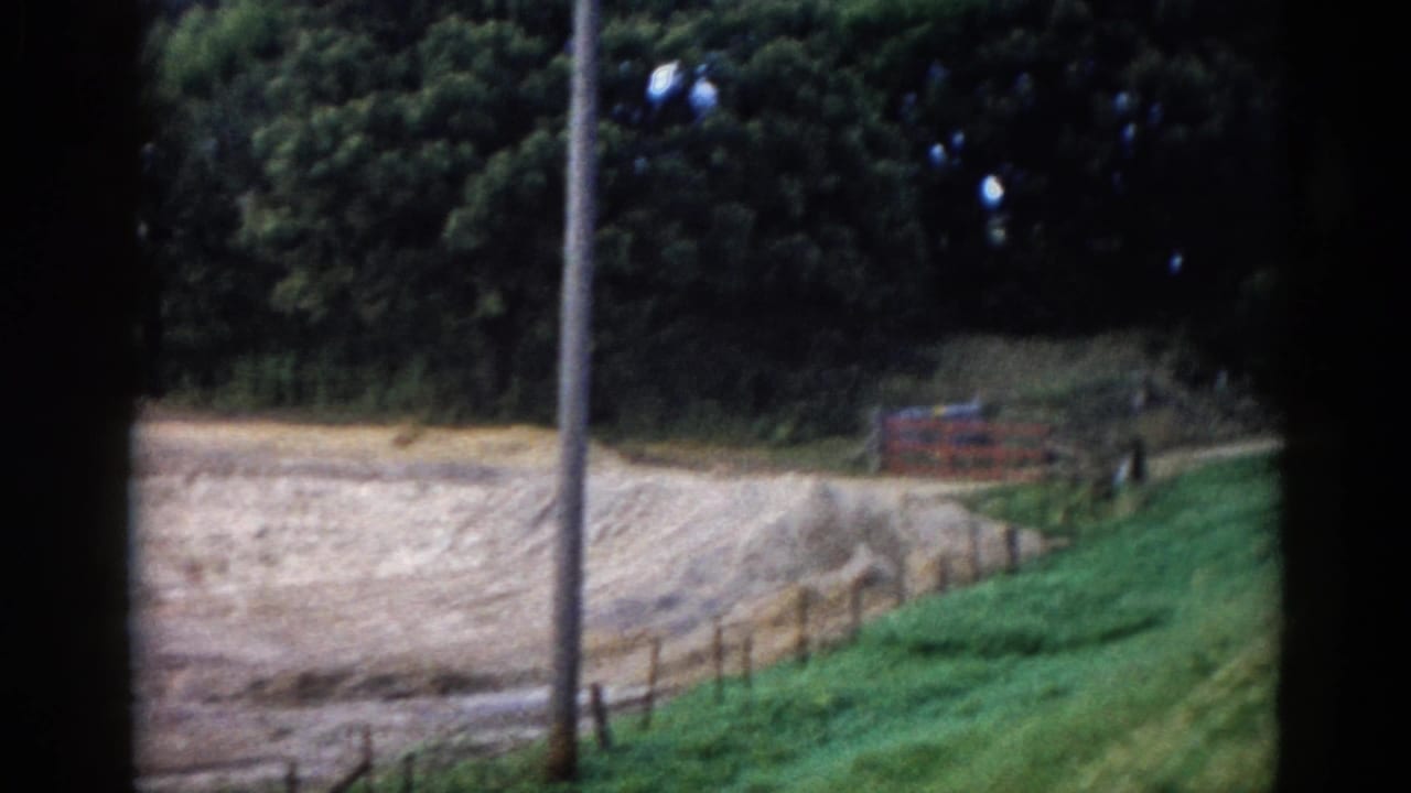 View from a moving vehicle of a grassy roadside embankment with a wooden post-and-rail fence, red barn structure, and a utility pole beside a rural road.