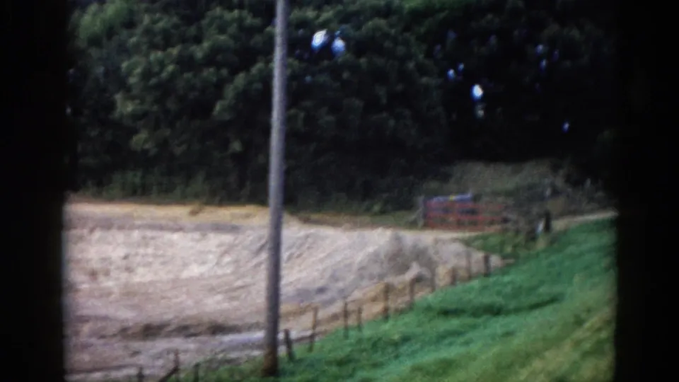 8mm home-movie frame from rural Illinois, circa 1962, showing a grassy embankment, wooden post-and-rail fence, red barn, and telephone pole along a country road.