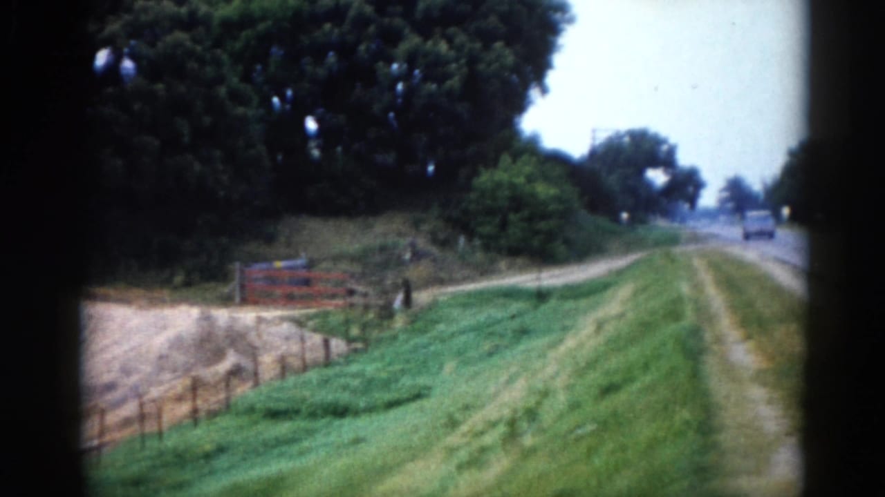 Rural two-lane road stretching ahead with a wooden fence, red barn, tree line, and a vehicle visible in the far distance.