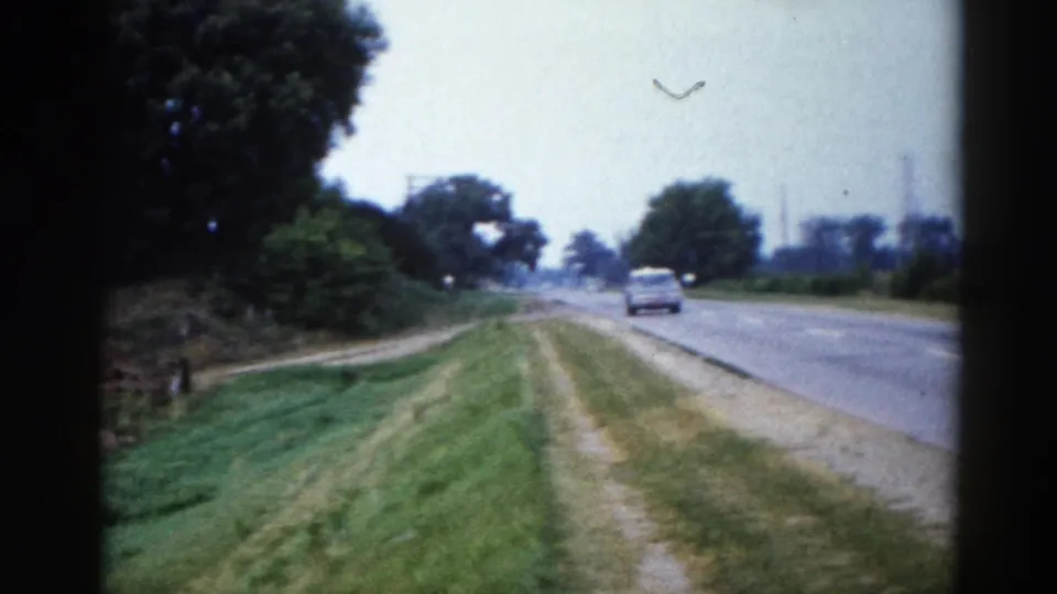 8mm road-trip footage from rural Illinois, circa 1962, showing a two-lane highway with mown grass verges, a distant light-coloured car, summer trees, and telephone poles.