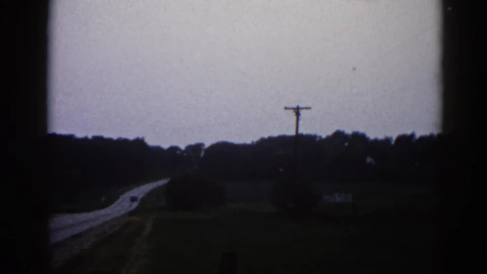 Underexposed 8mm frame, likely rural Illinois circa 1962, showing a dark flat landscape, a utility pole with crossarms, and a pale sky above a faintly visible receding road.