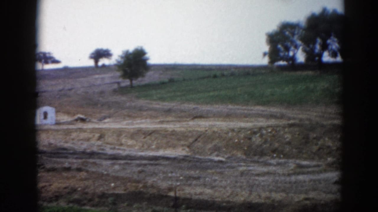 Rural hillside pan with graded dirt embankment, green slope, trees on ridge, and a small white structure at left edge, shot on 8mm film.