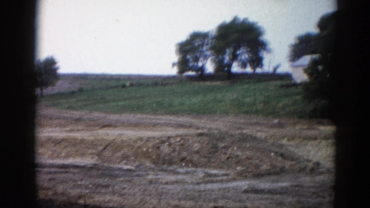 Continuing pan over a rural embankment with a green hillside, ridge trees, and a white building appearing at the right edge.