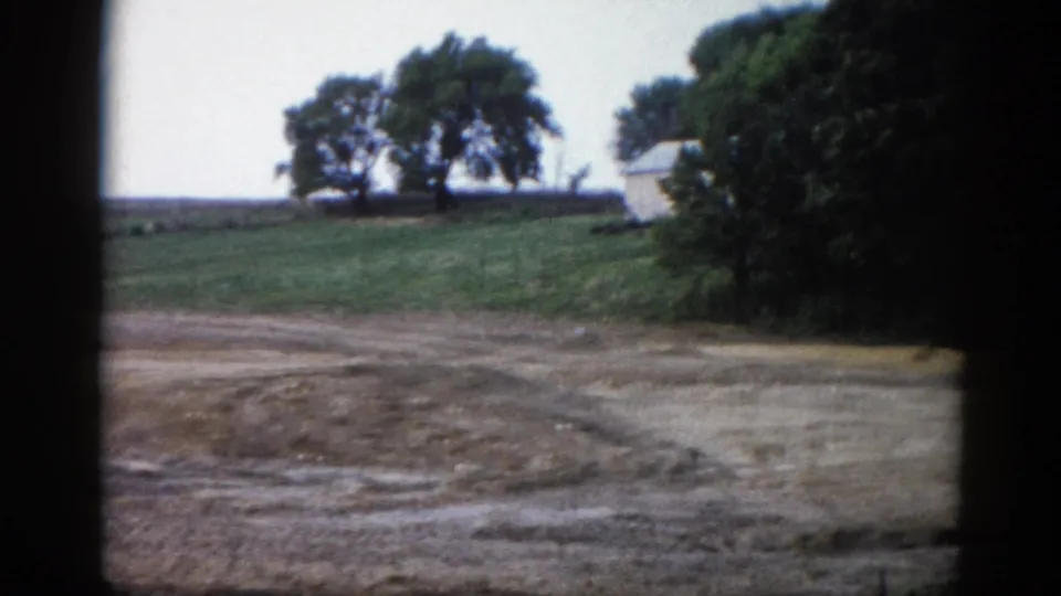 8mm home movie circa 1962 capturing an Illinois farmstead area with trees and two small white outbuildings visible through a panning shot.
