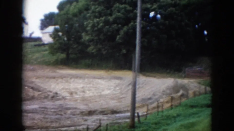 Final 8mm frame from a 1962 Illinois countryside pan, showing a utility pole, wooden fence rail, white house behind trees, and a dirt road curving through.