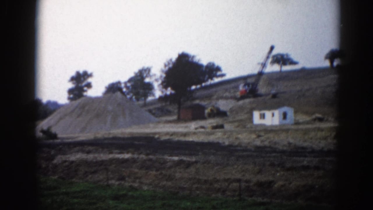 8mm home movie frame, c.1962, Illinois — small white site building and lattice-boom crane on a graded hillside with a large gravel spoil mound.
