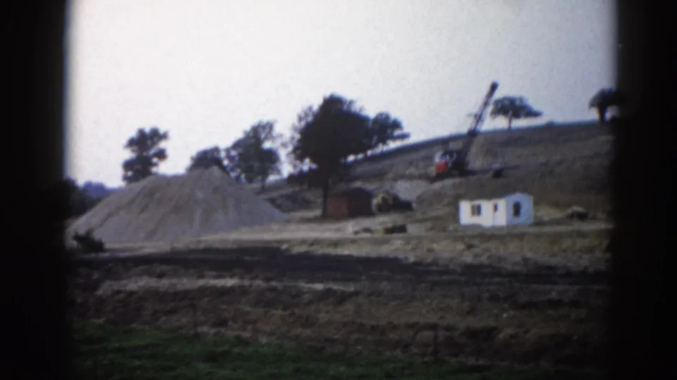 8mm home movie frame, c.1962, Illinois — small white site building and lattice-boom crane on a graded hillside with a large gravel spoil mound.