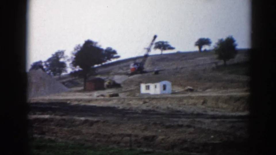 8mm frame, c.1962, Illinois — slow pan reveals white site structure and lattice-boom crane amid graded earth and large gravel mound.