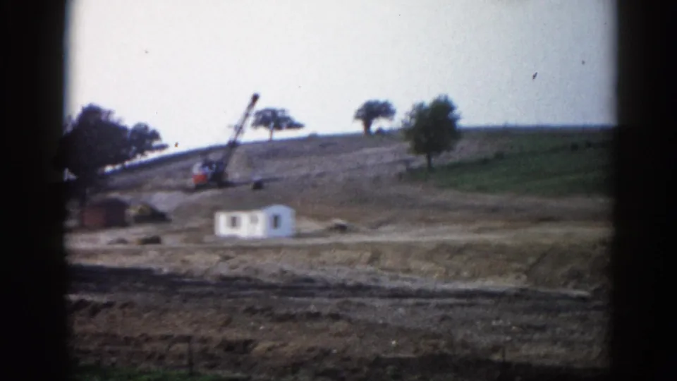 8mm frame, c.1962, Illinois — pan shifted to reveal green hillside; white building lower-left, crane centered on cleared earth.