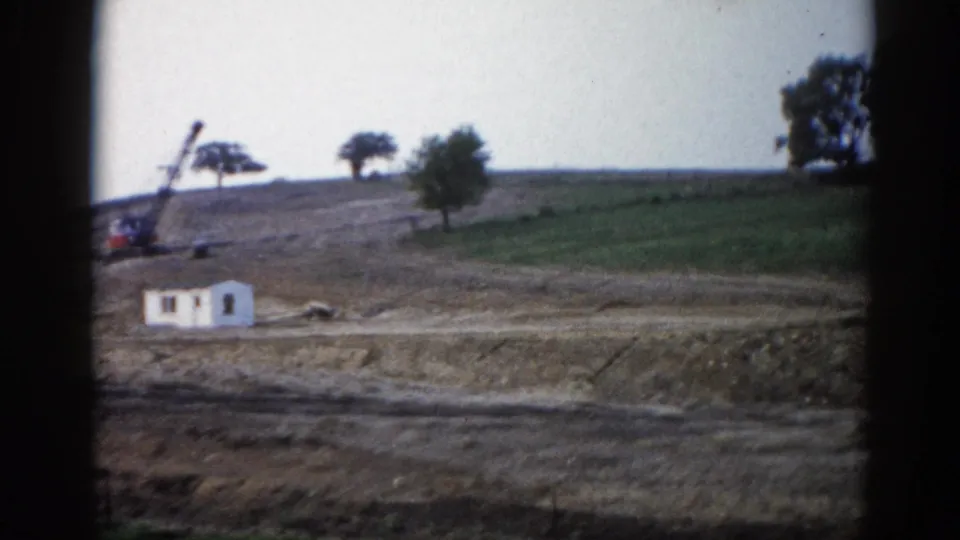 8mm frame, c.1962, Illinois — wide end-of-pan view; white structure left, crane right, open green landscape beyond cleared construction zone.