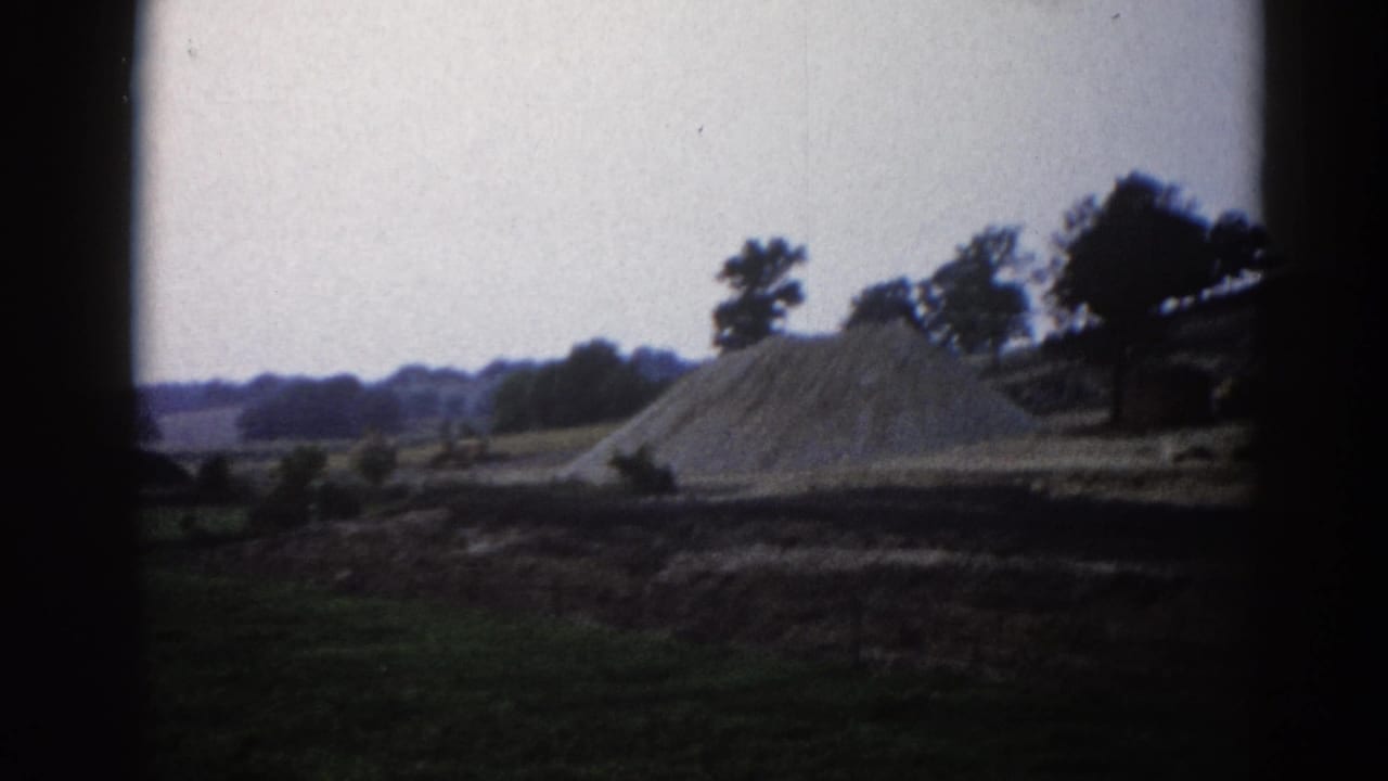 Large conical sand pile against a tree-lined horizon on a construction site, shot on 8mm film with visible edge vignetting.